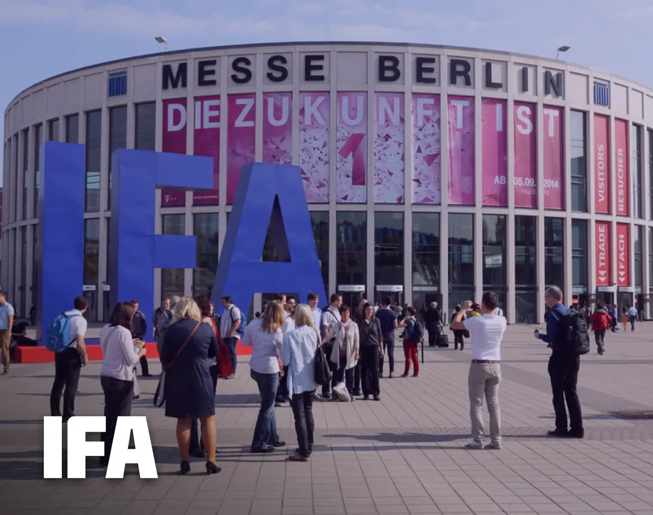 Large blue IFA letters in front of Messe Berlin building, with people gathering outside under sunny skies.