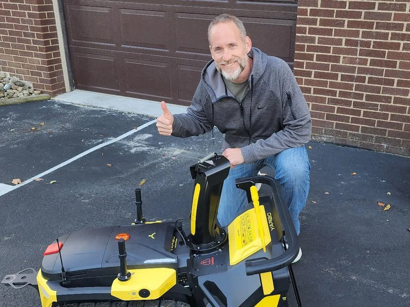 Man giving thumbs-up beside yellow and black robotic lawn mower in driveway, brick house background.