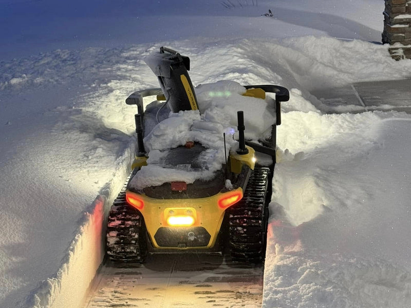 Yellow snow blower clearing deep snow at night, illuminated by bright lights, tracks visible in snow.