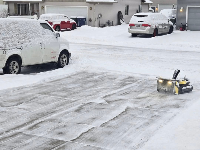 Snow-covered driveway with cleared path, yellow snow blower in foreground, vehicles and houses in background.