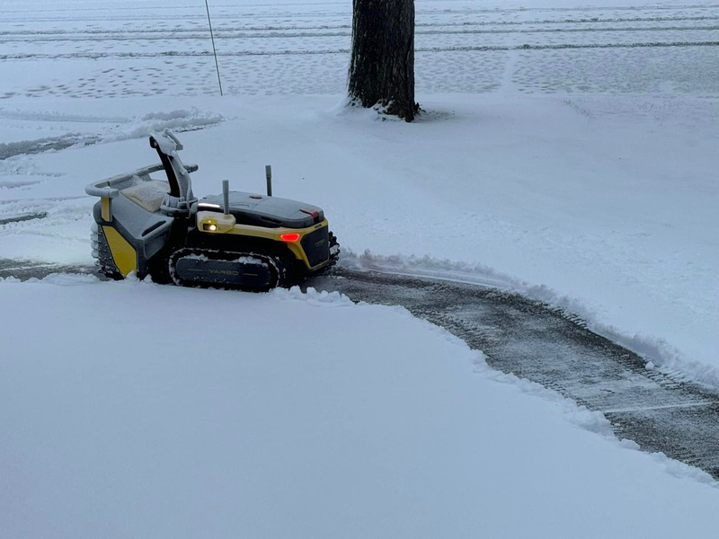 Yellow and black robotic lawn mower clearing snow from a driveway, with illuminated lights and a tree in the background.