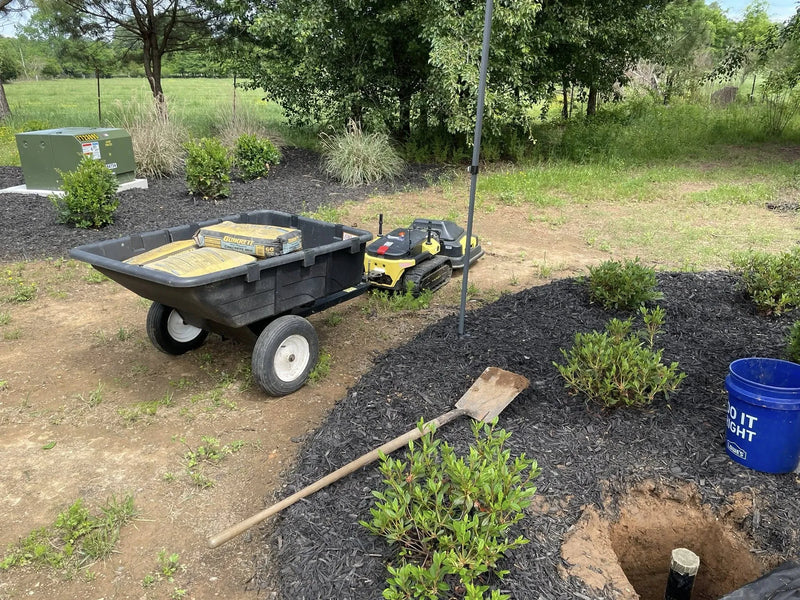 Black wheelbarrow with Quikrete bags, yellow robot, shovel, blue Lowe’s bucket, and mulched garden bed under trees.