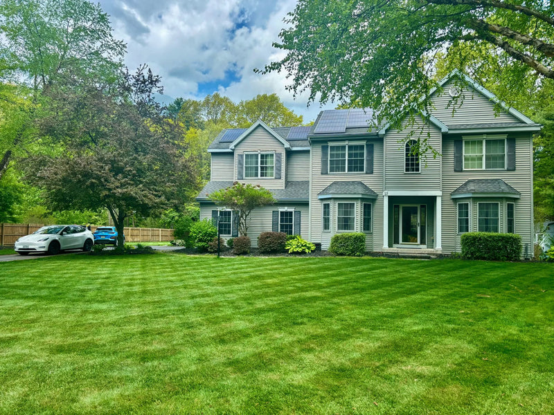Gray two-story house with solar panels, lush green lawn, white Tesla, and trees under partly cloudy sky.