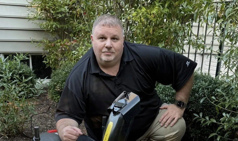 Man in black polo shirt and khaki pants kneels outdoors with garden tool, surrounded by green shrubs and house siding.