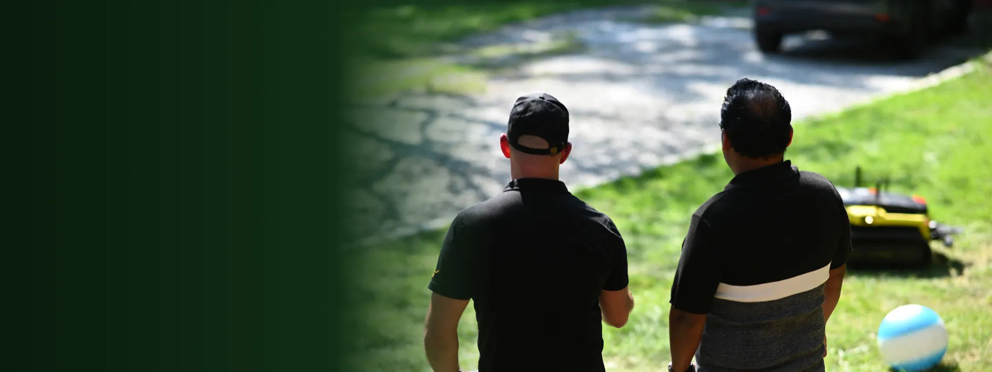 Two men in black shirts walk away on a sunny day, one wearing a cap, near a yellow lawnmower and blue-and-white ball on grass.